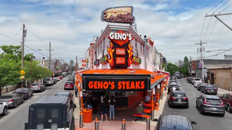 Aerial-view-of-Geno’s-Steaks,-a-bustling-cheesesteak-restaurant-with-vibrant-orange-signage-and-outdoor-seating,-located-on-a-busy-street