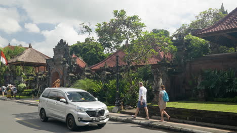 Man-and-woman-tourists-walking-along-sidewalk-in-front-of-Ubud-Palace,-opposite-traffic---panning-slow-motion