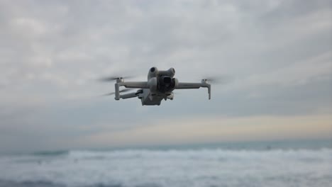 Isolated-drone-hovering-in-sky-over-beach-and-waves,-DJI-Mini-4-Pro