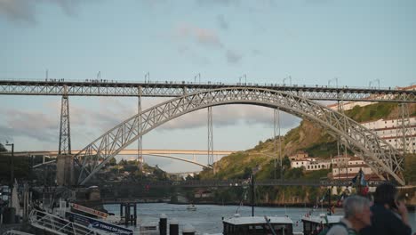 Dom-Luís-Bridge-spanning-the-Douro-River-in-Porto-at-sunset,-with-people-walking-and-boats-below