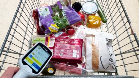 Customer-holding-a-Zebra-scan-as-you-shop-handheld-scanner-whilst-pushing-a-shopping-trolley-of-groceries-at-Tesco-supermarket-store