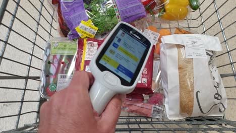 Person-with-Zebra-scan-as-you-shop-scanner-pricing-handheld-device-with-a-trolley-of-shopping-groceries-in-Tesco-supermarket-store