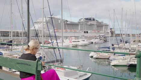 Woman-sit-on-wooden-bench-near-boat-dock-with-large-cruise-ship,-Madeira