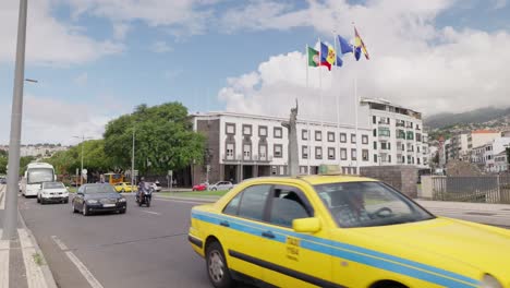 Vehicles-drive-on-main-street-of-Funchal-city,-flags-flutter-in-wind,-Madeira