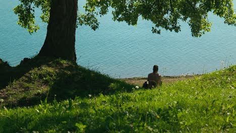 Person-sitting-by-the-shoreline-of-Jarun-Lake-in-Zagreb,-shaded-by-a-tree,-with-lush-grass-and-a-serene-lake-view