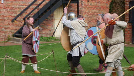 Mittelalterliche-Wikinger-Reenactors-In-Authentischen-Kostümen-Beteiligen-Sich-An-Einer-Nachgestellten-Schlacht,-Bei-Der-Sie-Mit-Speeren-Und-Schildern-In-Einem-Grasbewachsenen-Innenhof-Kämpfen