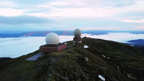 Two-radar-domes-sit-on-a-mountain-peak,-overlooking-a-sea-of-clouds-as-the-sun-rises