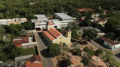 Panorámica-Aérea-De-La-Iglesia-De-San-Juan-Con-Techo-Amarillo-Y-Naranja-En-La-Isla-De-Margarita,-Venezuela,-Rodeada-De-árboles.