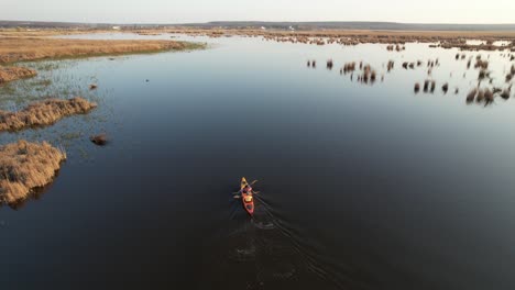 Two-people-kayaking-on-a-serene-lake-surrounded-by-reeds-and-vast-landscape-at-sunset