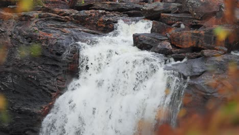 Amidst-the-Norwegian-autumn-forest,-a-small-waterfall-gracefully-cascades-over-dark-rocks,-enveloping-the-scene-in-a-serene-ambiance-of-natural-beauty