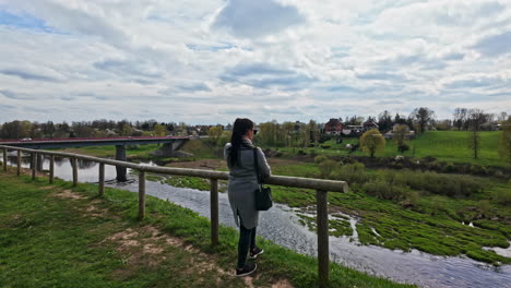 Young-woman-with-black-hair-looking-at-small-river-water-stream-near-bridge