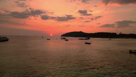 El-Sol-Se-Pone-Sobre-El-Océano-En-Calma,-Proyectando-Un-Cálido-Resplandor-Sobre-Los-Barcos-Y-Una-Isla-En-La-Distancia,-Pintando-El-Cielo-En-Tonos-Naranja-Y-Rosa