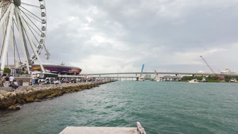 Bayside-view-of-Miami-with-ferris-wheel,-a-bridge,-boats,-and-construction-cranes-on-a-cloudy-day