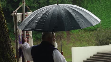 A-senior-man-holding-an-umbrella-stands-watching-the-rain-fall,-creating-a-peaceful-and-contemplative-scene