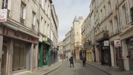 People-walking-and-cycling-on-a-historic-street-in-La-Rochelle,-France-with-shops-and-an-ancient-tower