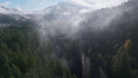 Wispy-fog-or-clouds-reveal-river-in-epic-valley-forest-with-evergreens-rolling-along-mountains