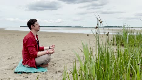 Hübscher-Mann-Sitzt-Auf-Einer-Yogamatte-Und-Beginnt-Bei-Sonnigem-Wetter-Am-Strand-An-Der-Ostsee-Zu-Meditieren,-4k,-Mittlere-Aufnahme,-30 fps