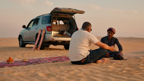 Dos-Hombres-Sentados-Junto-A-Un-Coche-En-El-Desierto-Del-Sahara,-Con-Tablas-De-Arena-Y-Un-Picnic