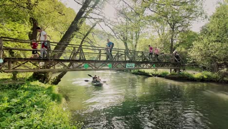 Families-walk-over-rustic-wooden-footbridge-as-canoe-couple-paddle-on-river