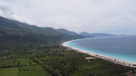 Aerial-view-of-Borsh-Beach-in-Albania,-showcasing-its-expansive-shoreline,-turquoise-waters,-and-lush-green-surroundings-under-a-partly-cloudy-sky