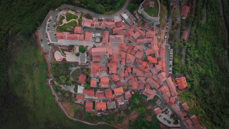Top-down-aerial-small-town-on-top-of-mountain-surrounded-by-trees,-Italy