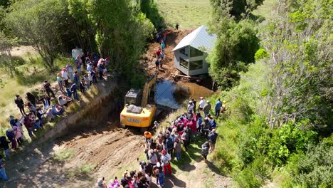 Vista-Aérea-De-Una-Excavadora-Y-Una-Gran-Comunidad-Local-Ayudando-A-Trasladar-Una-Casa-Al-Otro-Lado-Del-Río-En-Chiloé,-Chile.