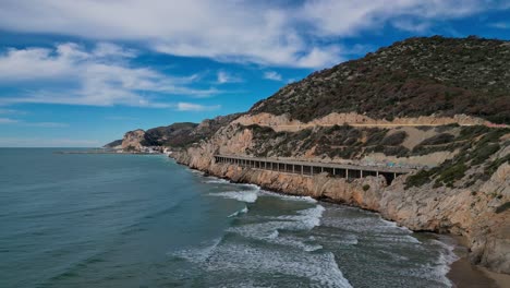 Vista-Panorámica-De-La-Costa-De-Port-Ginesta-Con-Montañas-Y-Olas-Bajo-Un-Cielo-Azul-Brillante