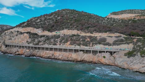 Vista-Panorámica-De-La-Carretera-Costera-A-Lo-Largo-De-Los-Acantilados-De-Port-Ginesta-Con-Aguas-Turquesas-Y-Olas.