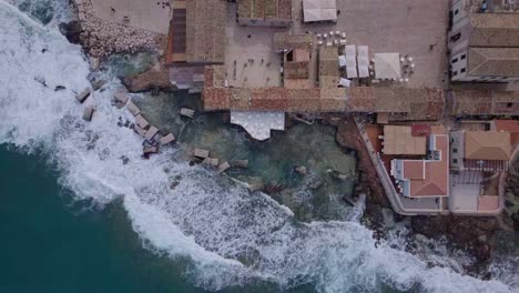 Aerial-Top-down-waves-crashing-into-a-restaurant-deck,-Marzamemi,-Sicily