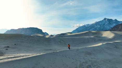 A-solitary-figure-is-seen-running-through-expansive-sand-dunes-with-majestic,-snow-capped-mountains-in-the-distance