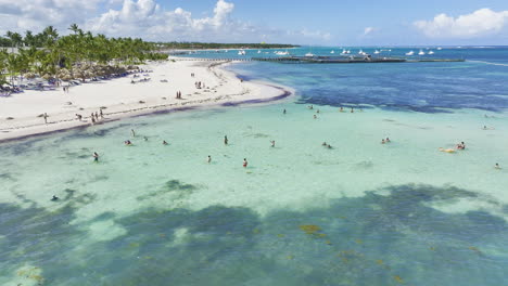 Panning-from-the-right-to-the-left-side-of-the-frame-and-slowly-approaching-the-beachfront-where-tourists-are-swimming-and-sunbathing-by-the-sea-located-in-Dominican-Republic-in-the-Caribbean