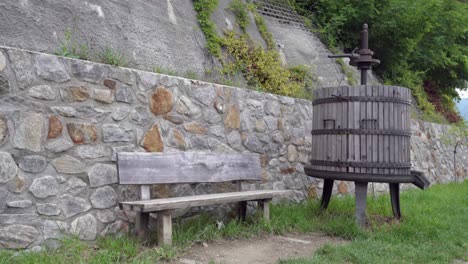 A-wooden-bench-and-a-vintage-grape-press-in-front-of-a-stone-wall-on-the-irrigation-channel-path-in-Algund---Lagundo,-South-Tyrol,-Italy