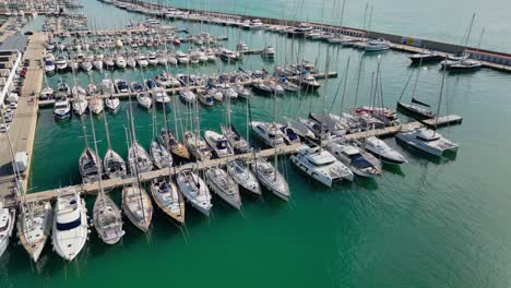 The-coast-and-port-of-ginesta-in-barcelona-with-docked-boats-in-clear-blue-water,-aerial-view