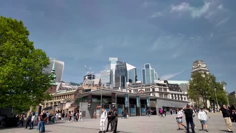 Eine-Geschäftige-Stadtumgebung-Mit-Einem-Platz-Mit-Spazierenden-Menschen-Und-Modernen-Wolkenkratzern-Im-Hintergrund-Unter-Einem-Klaren-Blauen-Himmel