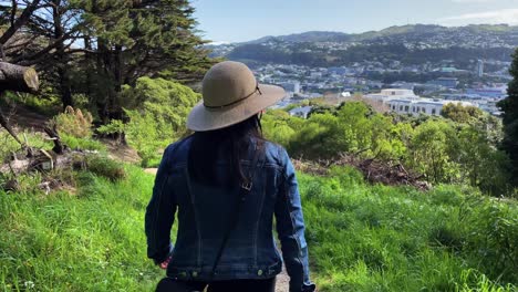 A-woman-with-black-hair-wearing-a-sun-hat-and-blue-denim-jacket-walks-down-a-dirt-hiking-trail-through-a-forest-towards-the-view-of-the-city-of-Wellington,-New-Zealand