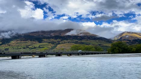 Un-Solo-Automóvil-Cruza-El-Puente-Del-Río-Reese-En-Glenorchy-Paradise-Road-En-Nueva-Zelanda-Frente-A-Grandes-Colinas-En-Un-Día-Nublado