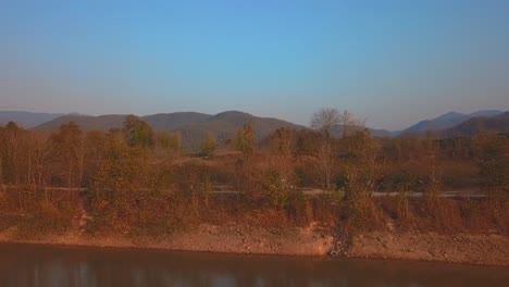 The-drone-shot-captures-a-car-driving-along-the-lakeshore,-set-against-a-backdrop-of-mountains-during-sunset