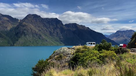 An-RV-camper-is-parked-alone-at-the-edge-of-a-cliff-overlooking-Lake-Whatipu-and-a-mountain-range-on-a-beautiful-summer-day-in-New-Zealand