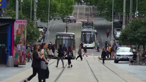 Slow-motion-shot-of-hustle-and-bustle-of-Melbourne-urban-lifestyle,-tram-running-along-Bourke-street,-passing-through-a-bustling-city-with-pedestrians-crossing-and-commuters-waiting-at-the-tram-stop