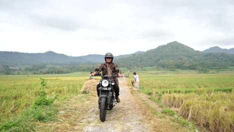 Biker-guy-driving-a-motorbike-in-a-rice-field-area-with-mountain-views-with-rural-atmosphere