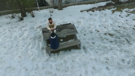 Drone-flies-slowly-over-people-sitting-on-a-bench-and-a-table---there-is-snow-on-the-ground-and-the-people-are-warmly-dressed