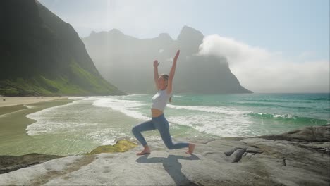 Woman-doing-morning-sun-salutation-pose-in-early-morning-on-Norway-coast,-side-view