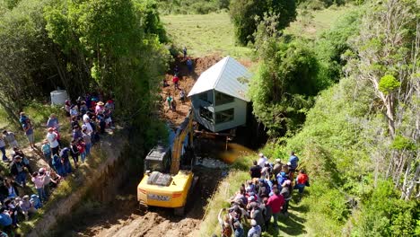 Vista-Aérea-De-La-Excavadora-Y-Una-Gran-Comunidad-Local-Ayudando-A-Trasladar-Una-Casa-A-Través-Del-Río-En-Chiloé,-Chile.