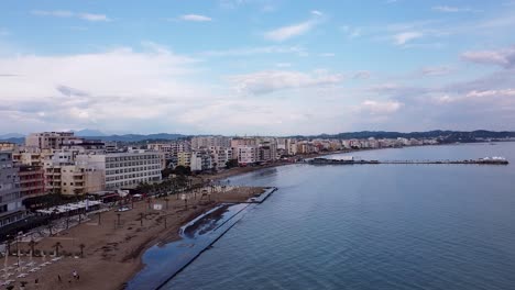 Aerial-view-of-Durrës,-Albania,-featuring-the-city's-beachfront,-coastal-buildings,-and-calm-blue-waters-under-a-partly-cloudy-sky