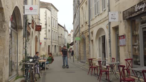 Tilt-down-shot-of-locals-passing-by-chair-tables-arranged-outside-a-café-in-a-narrow-lane-in-La-Rochelle,-France-on-a-cloudy-day