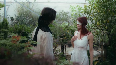 Cinematic-medium-shot-video-of-couple-blindfolded-approaching-slowly-in-greenhouse-surrounded-by-plants-and-flowers