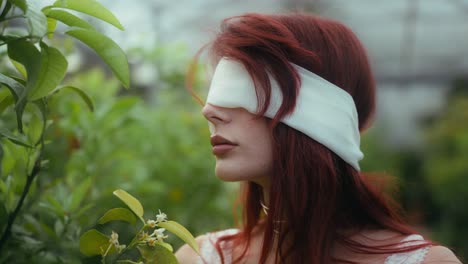 cinematic-circle-pan-footage-of-a-young-woman-in-a-white-dress-and-blindfolded-looking-around-in-a-greenhouse-plant