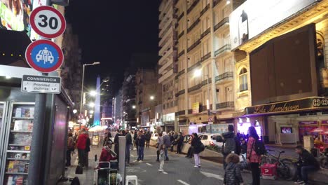 People-walk-at-Corrientes-avenue-famous-city-center-with-obelisk-background-at-Buenos-Aires-city-Argentina-night,-illuminated-stores-and-buildings