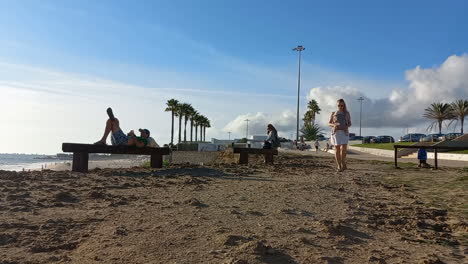 Sea-wall-next-to-the-beach-with-benches-to-look-at-the-sea-and-relax,-young-woman-passing-by-with-a-baby-carrier-backpack,-blue-sky-with-gray-clouds,-relaxing-place