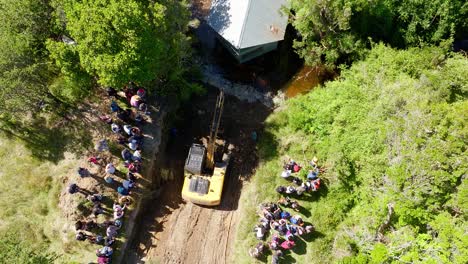 Aerial-View-Of-Excavator-And-Large-Local-Community-Helping-To-Move-House-Across-River-In-Chiloe,-Chile